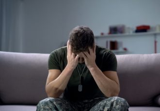 A veteran in uniform sits on a couch with his head in his hands, depicting emotional strain and the need for support.